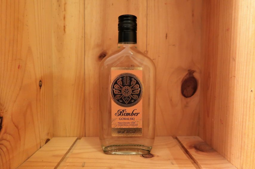 Eine transparente Glasflasche mit schwarzem Deckel und gold-orangenem Etikett steht in einer Holzkiste.