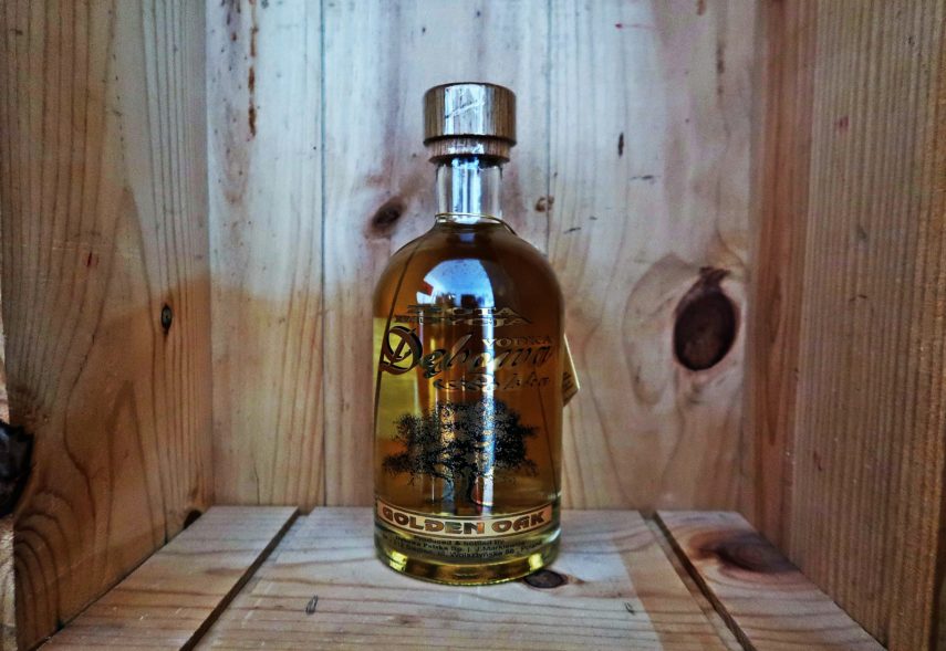 Eine Glasflasche mit goldener Schrift und goldfarbenen Wodka. Sie steht in einer Holzkiste.