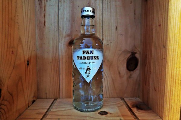 Eine transparente Glasflasche mit silbernem Deckel und Wodka als Inhalt steht in einer Holzkiste. Das weiße Etikett wird von einer polnischen Literaturfigur geziert.