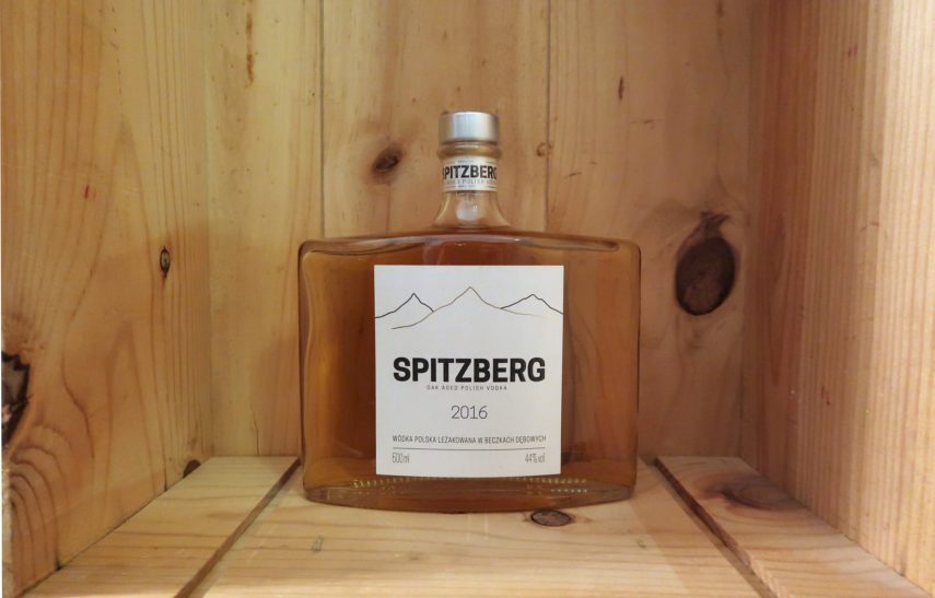 Eine transparente Glasflasche mit silbernen Verschluss und weiß-grauem Etikett steht in einer Holzkiste.