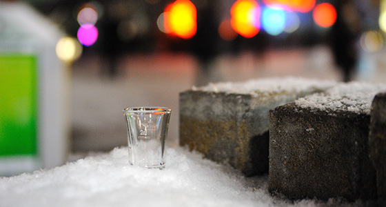Ein Schnapsglas steht im Schnee. Daneben liegen ein paar Steine und im Hintergund sieht man bunte Lichter.